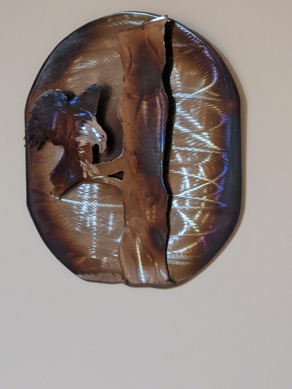 Oval heat-tinted steel art of an eagle landing on a pine